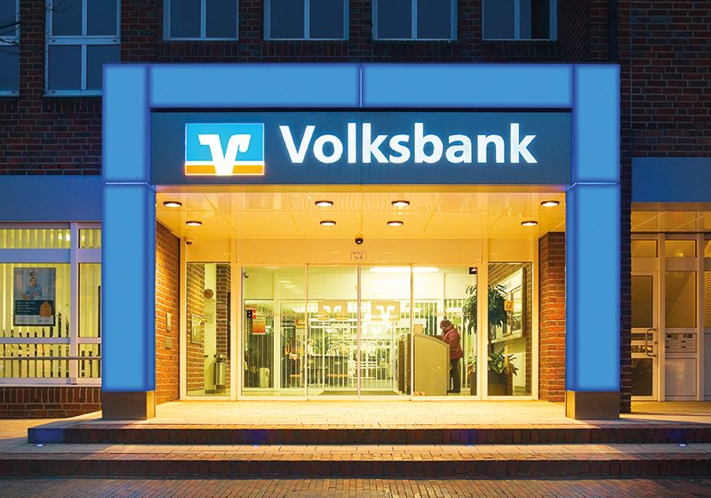 Beleuchtetes Eingangsportal einer Volksbank-Filiale in der Abenddämmerung, Logo und Schriftzug leuchten über dem Eingang
